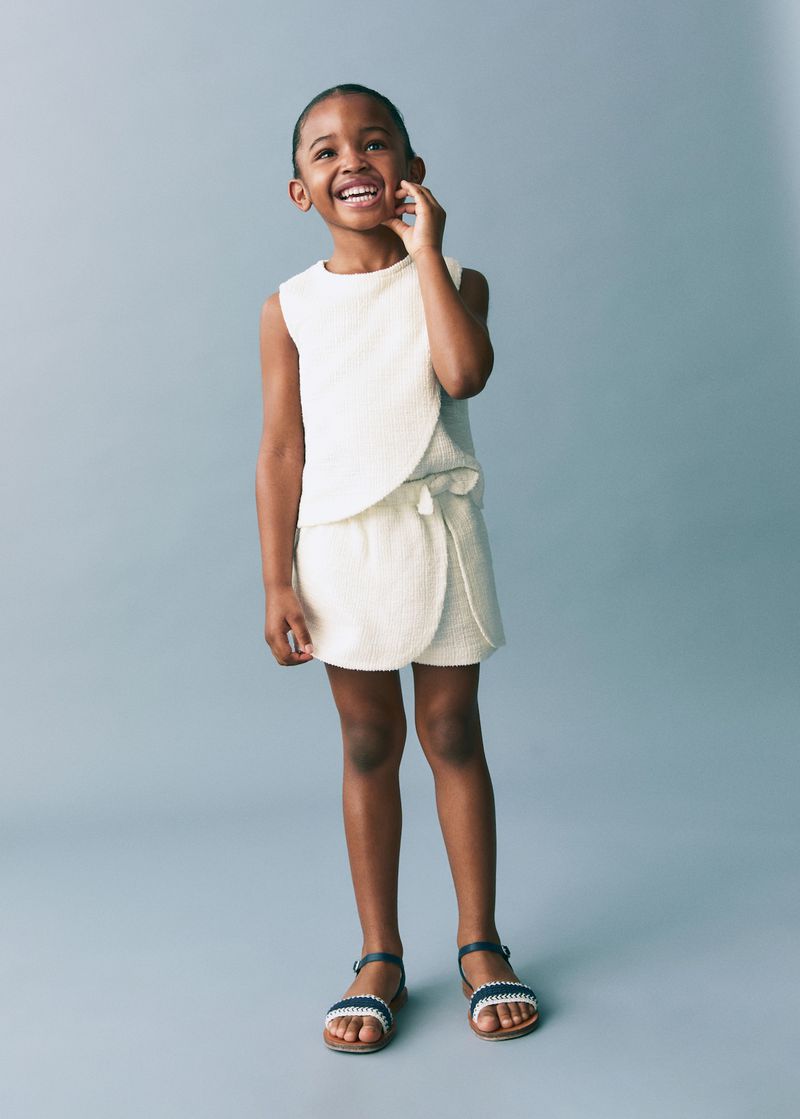 Smiling Black toddler girl wearing a textured white sleeveless top and shorts against a neutral grey studio backdrop.