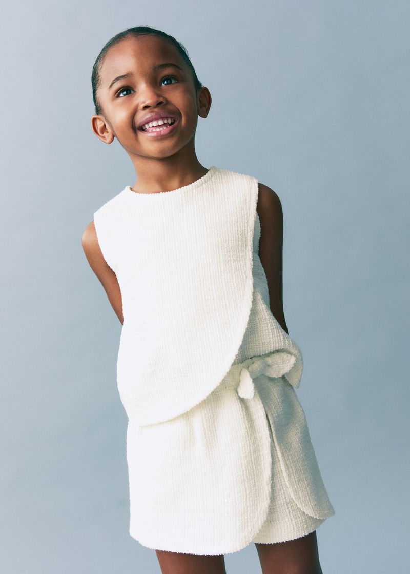 Portrait of a smiling young black girl wearing a sleeveless cream-colored textured top and wrap skirt, against a pale blue background.