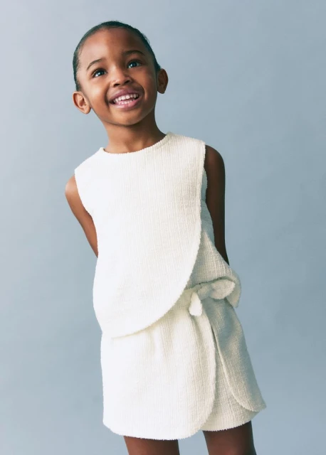 Portrait of a smiling young black girl wearing a sleeveless cream-colored textured top and wrap skirt, against a pale blue background.