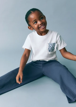 Smiling young Black girl wearing a white t-shirt and wide blue pants, sitting on a light blue studio backdrop, looking up.