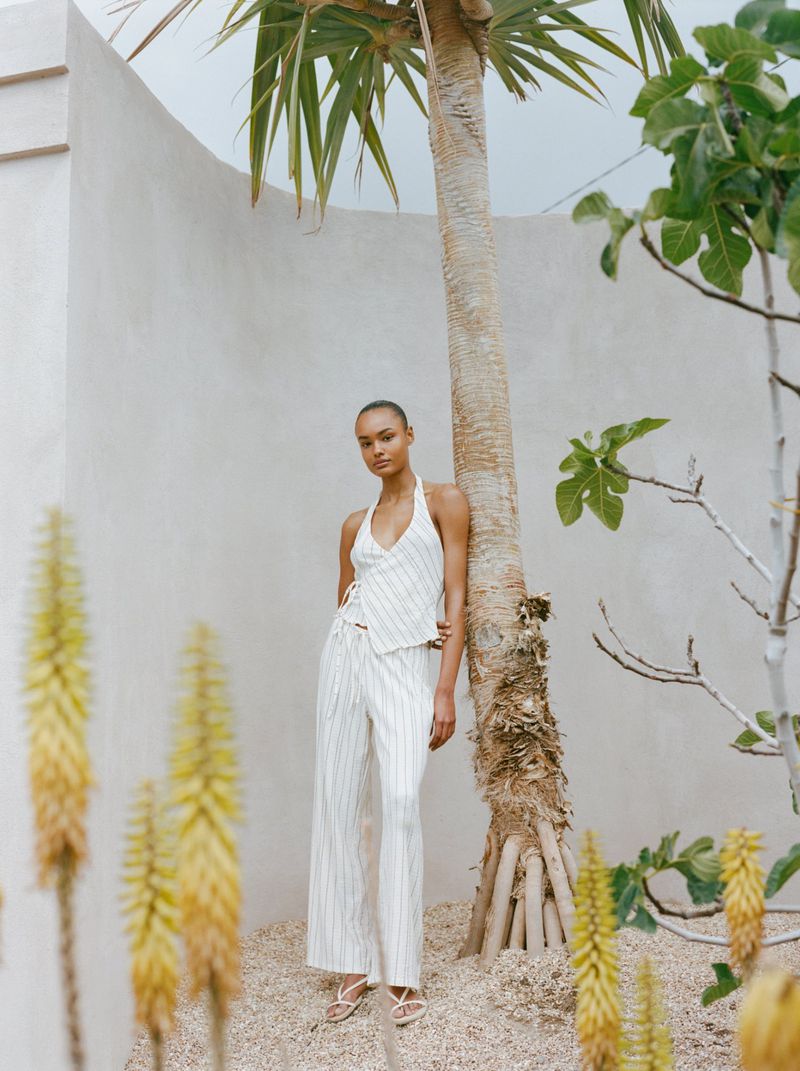 Woman in a halter top and pants poses next to a palm tree. She stands on a gravel surface in front of a textured white wall.