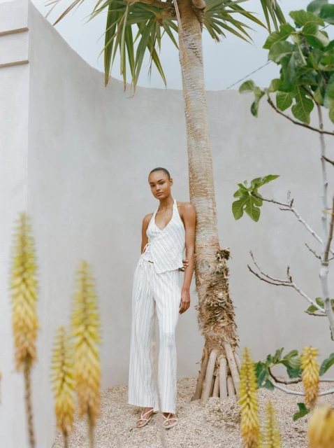 Woman in a halter top and pants poses next to a palm tree. She stands on a gravel surface in front of a textured white wall.