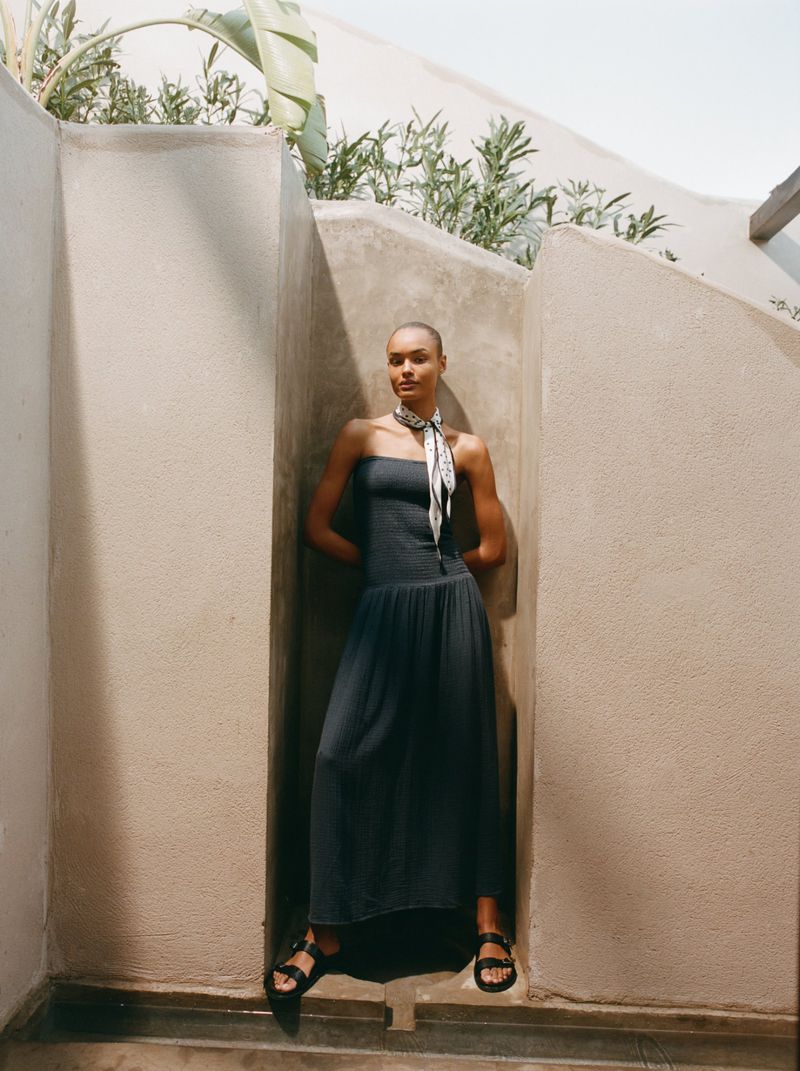 Adult female model wears a strapless dress and sandals, posing in a nook outdoors with green plants and soft, natural light. Fashion editorial.