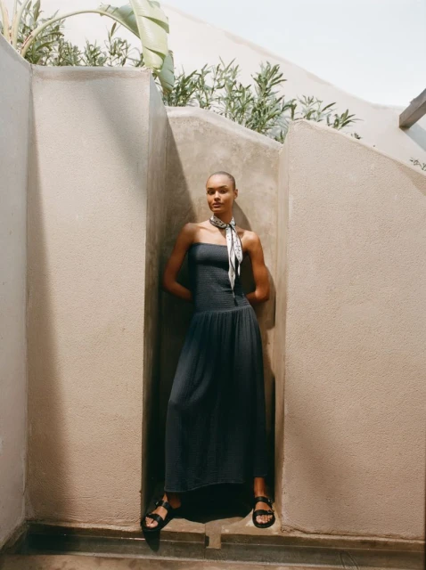 Adult female model wears a strapless dress and sandals, posing in a nook outdoors with green plants and soft, natural light. Fashion editorial.