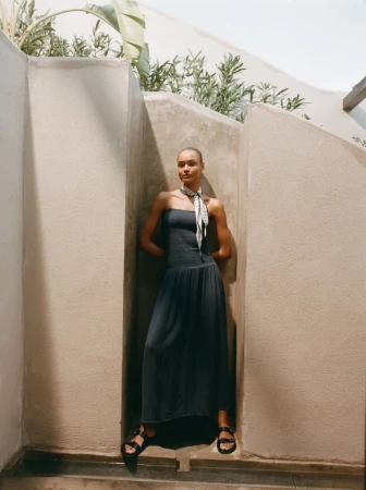 Adult female model wears a strapless dress and sandals, posing in a nook outdoors with green plants and soft, natural light. Fashion editorial.