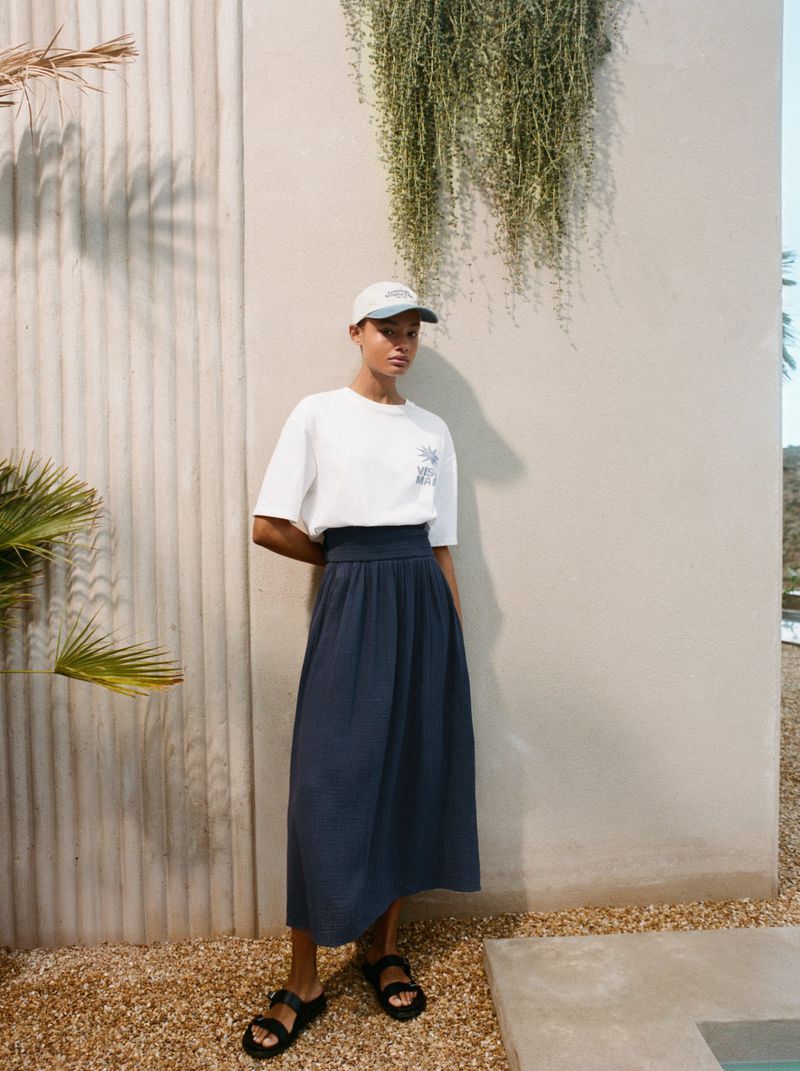 A young woman in a white t-shirt and blue skirt stands in front of a stucco wall, wearing sandals and a baseball cap in an outdoor setting.