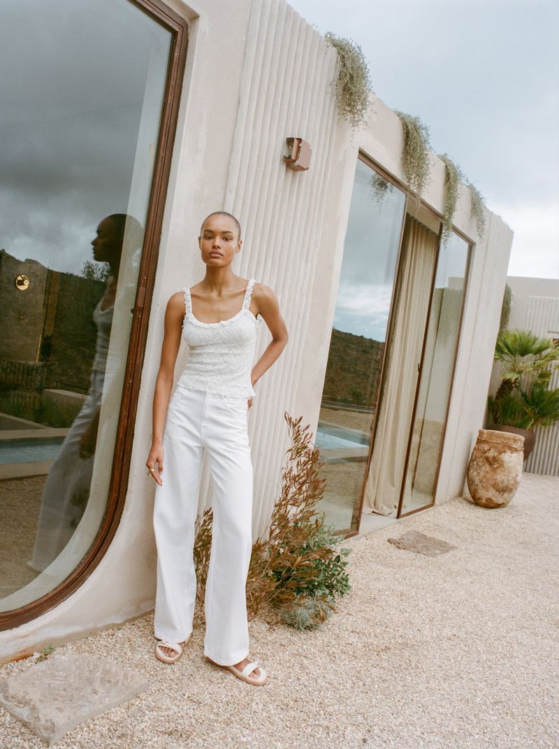 A woman with a shaved head wears white pants and top outdoors in front of an angular white building with potted plants.
