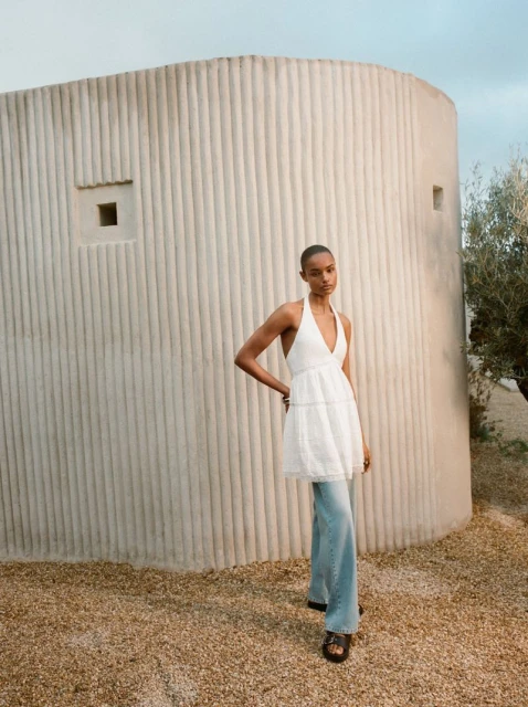 An adult female model is posing outdoors wearing a white halter top, loose-fitting blue jeans, and sandals near a modern adobe structure.