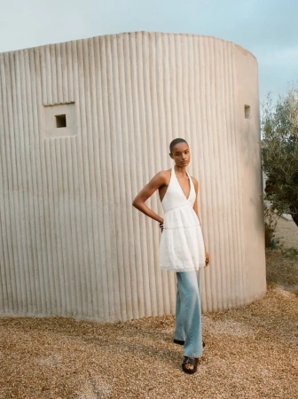 An adult female model is posing outdoors wearing a white halter top, loose-fitting blue jeans, and sandals near a modern adobe structure.