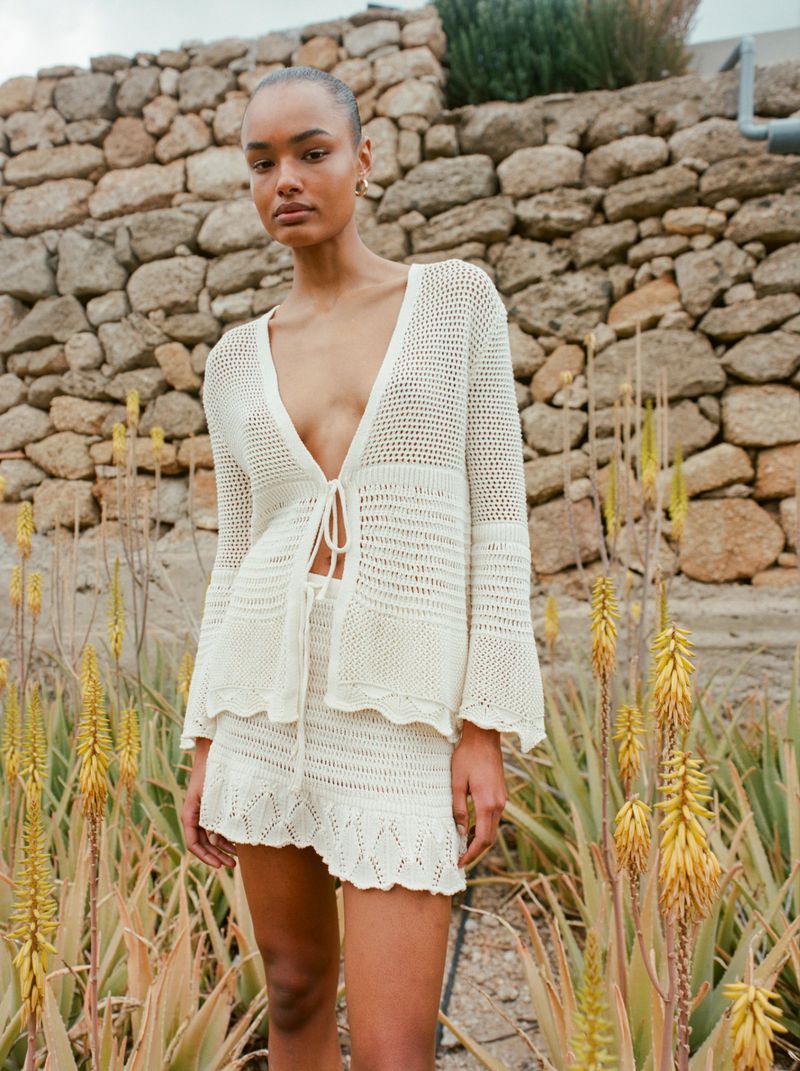 Woman in crochet knit two-piece set poses outdoors against a stone wall and natural greenery, showcasing fashion and style in natural light.