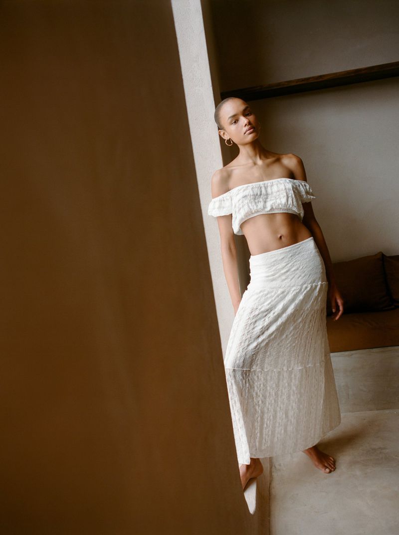 Woman in white off-the-shoulder top and long skirt posing near a wall, soft natural light, minimalist fashion, neutral tones.