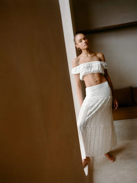 Woman in white off-the-shoulder top and long skirt posing near a wall, soft natural light, minimalist fashion, neutral tones.