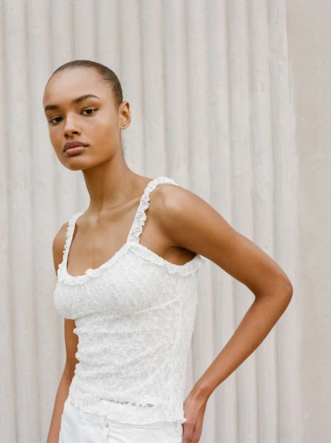 Young woman poses in a white lace tank top and white pants against a textured off-white background in a studio shot.