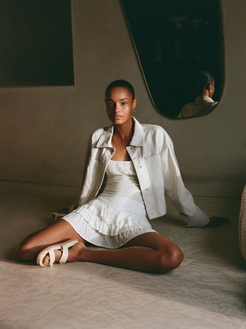 A young adult woman in a white dress and jacket is seated on the floor with a mirror in the background in an indoor setting.