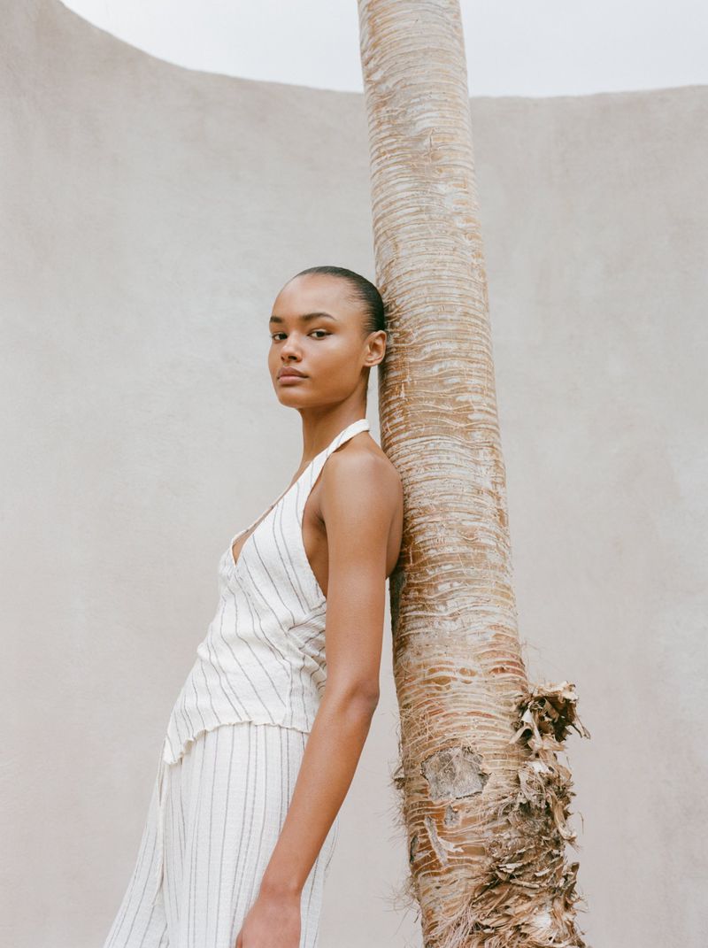 A slender woman with dark hair poses next to a textured palm tree trunk, wearing a cream-colored striped halter top and matching pants.