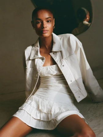 Model wearing a white denim jacket and matching white mini dress seated indoors under natural light