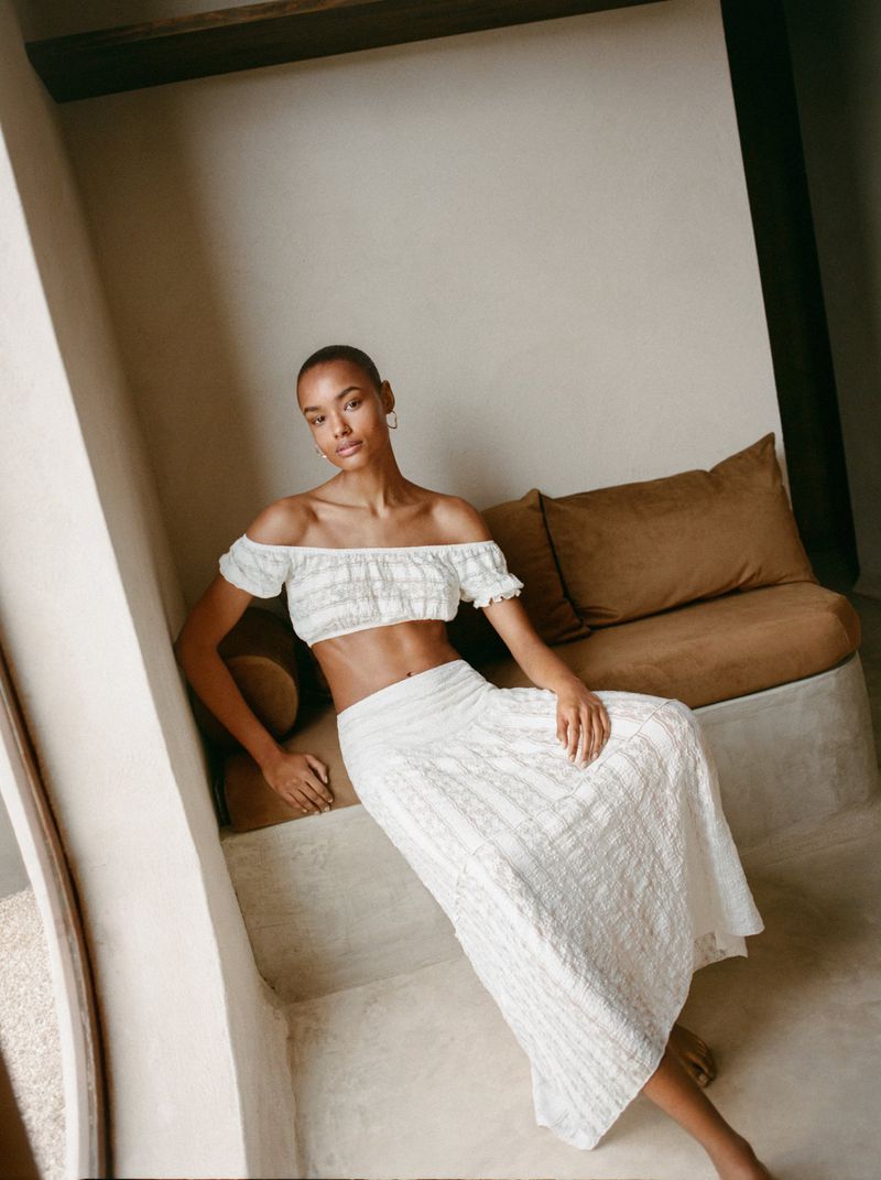 Woman in white off-the-shoulder crop top and maxi skirt poses indoors on a built-in bench with tan cushions in soft natural light.