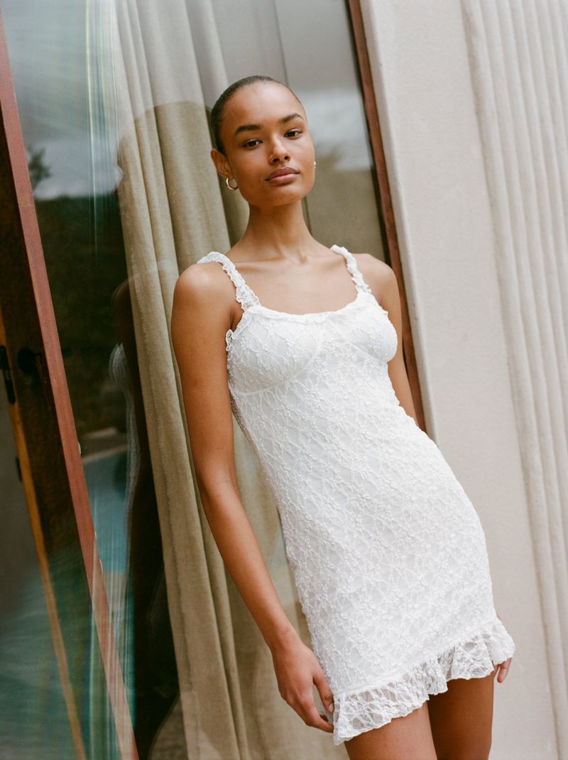 A woman in a white lace dress poses in front of a window, showcasing the dress's intricate details and her elegant style.