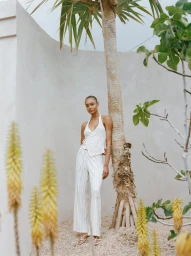 Model in white halter top and matching trousers leaning against a palm tree outdoors by a light stucco wall
