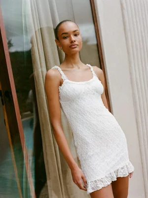 Model wearing a white lace slip dress standing against a light wall and curtained window for Pull and Bear.