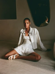 Model seated on the floor wearing a white denim jacket and matching mini dress with sandals in an indoor setting