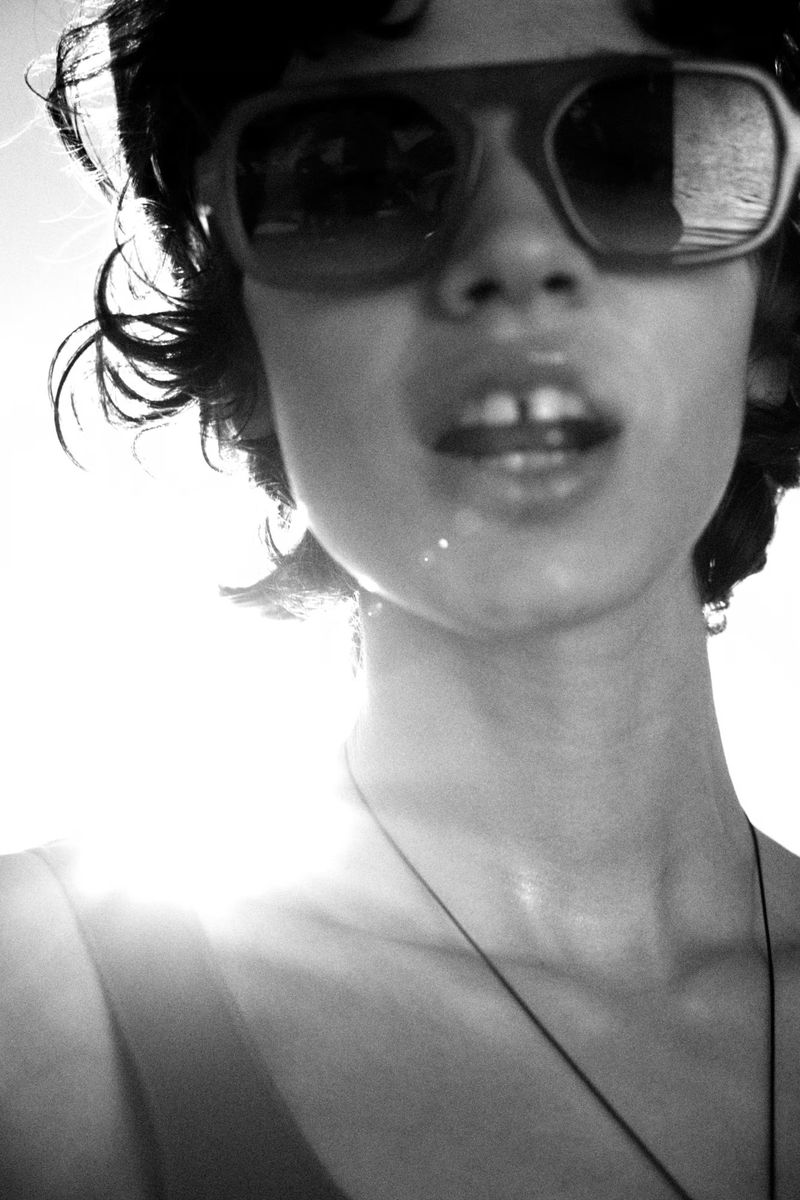 Black and white portrait of a young adult woman wearing large sunglasses, captured with sunlight creating lens flare in the background.