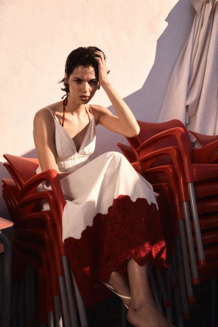 Young woman in white dress with red trim poses while seated on a stack of red chairs against a white wall with light shadows.
