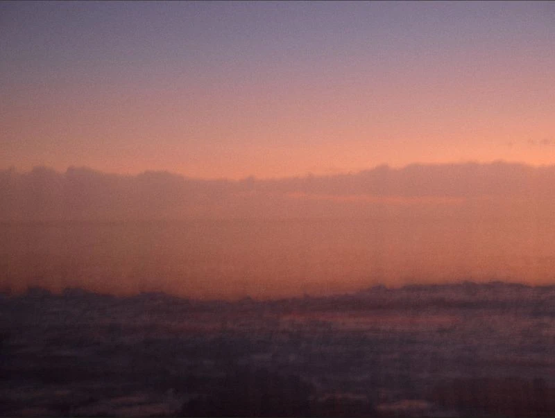 Sunrise or sunset sky with purple and orange hues over soft, low clouds. The scene has a hazy, atmospheric quality.