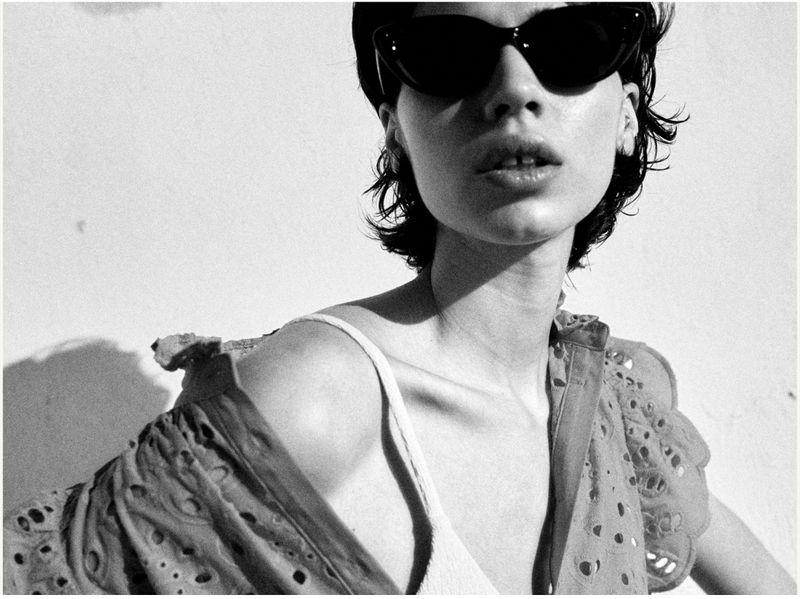 Monochrome portrait of an adult woman with dark sunglasses and short hair, wearing an open embroidered shirt with a white top.