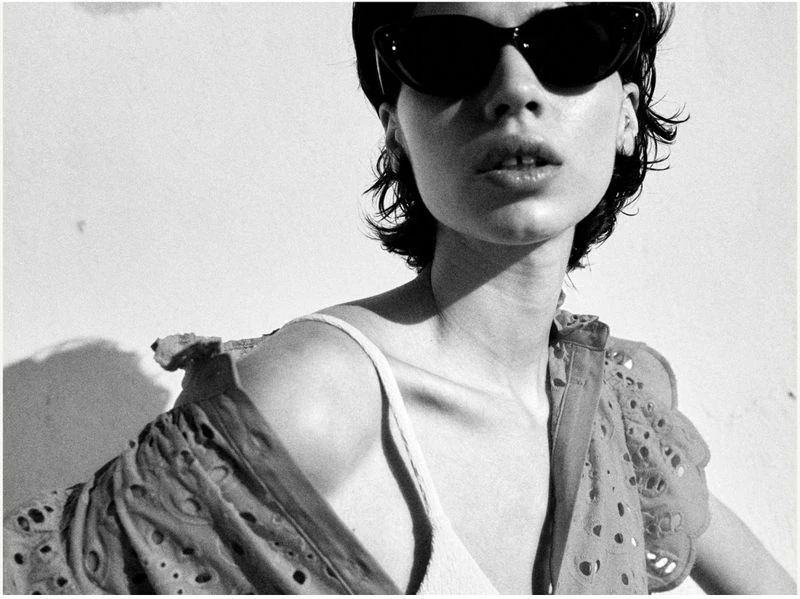 Monochrome portrait of an adult woman with dark sunglasses and short hair, wearing an open embroidered shirt with a white top.