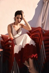 Model wearing a white dress with red hem seated amongst stacked red chairs outdoors under bright sunlight.