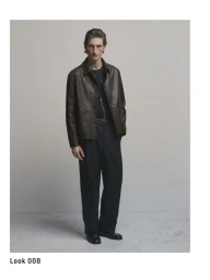 Full-length portrait of a male model wearing a dark leather jacket and trousers against a neutral grey studio background.