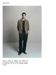 Model standing against a neutral grey studio background wearing a camel blazer over a black t-shirt and dark trousers.