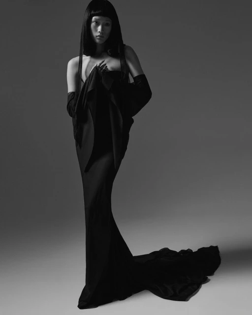 Asian woman in long black gown and opera gloves poses in studio. Fashion shot in monochrome with soft lighting and dark, straight hair.