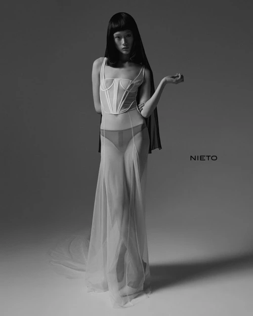 Black-and-white fashion shot of a slender adult woman in sheer white dress with bustier posing against a neutral grey backdrop in studio lighting.