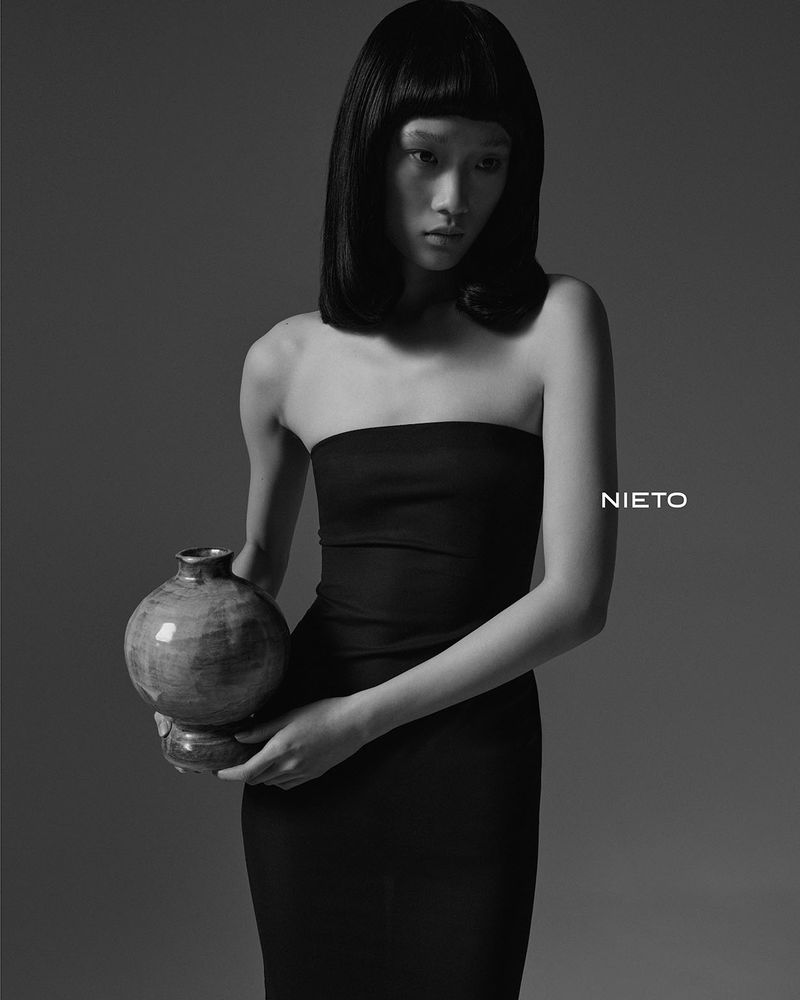 Studio shot of a slender female in a black dress holding a ceramic vase, showcasing dark hair and minimalist fashion against a grey backdrop.
