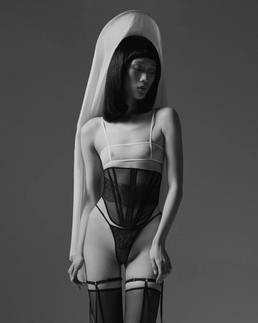 Black-and-white studio shot of a slender, posed Asian woman in lingerie and a sheer head covering, set against a grey backdrop.