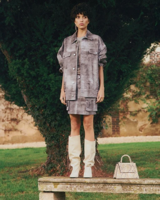 An adult female poses standing on a stone bench in an outdoor setting, wearing a matching grey patterned skirt and jacket.