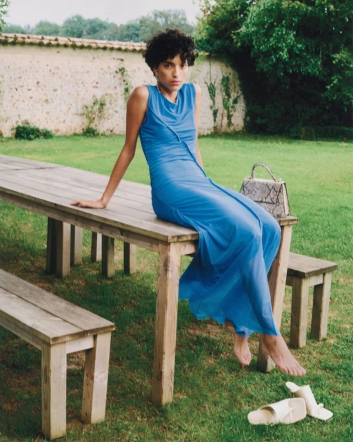 A woman in a blue dress sits on a wooden table outdoors on a lawn. A snakeskin purse and sandals rest on the grass.