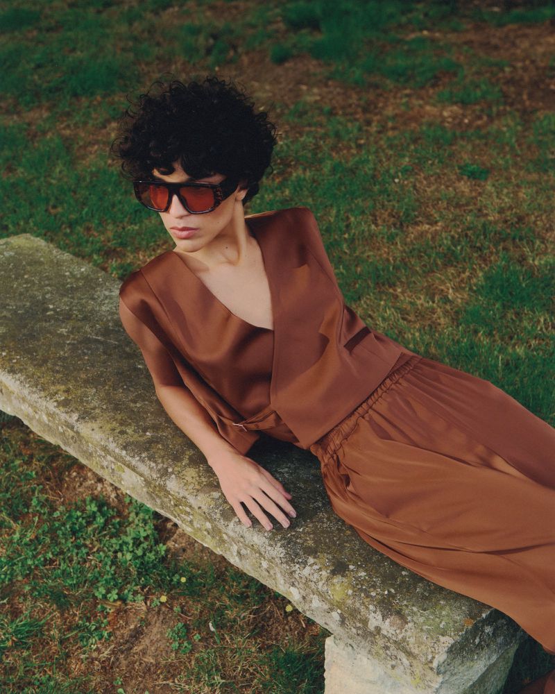 Woman in stylish sunglasses and brown outfit lounges on stone bench amidst green grass, exuding relaxed fashion in outdoor setting.