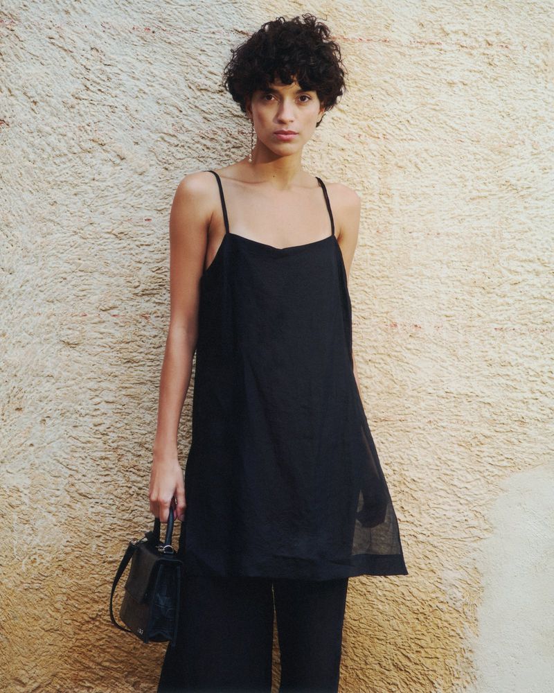 Fashionable woman in black top and wide-leg pants poses against a textured wall, carrying a small black handbag.
