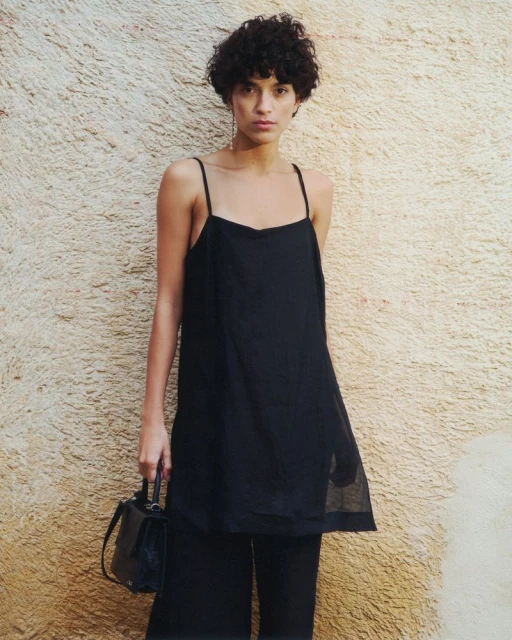 Fashionable woman in black top and wide-leg pants poses against a textured wall, carrying a small black handbag.