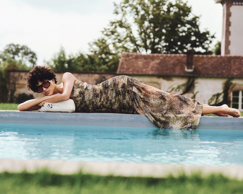 Woman in patterned dress lounges poolside on white towel wearing sunglasses with building and trees in background on sunny day.