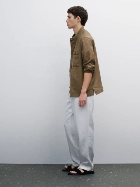 A man stands in profile, wearing an olive-green, long-sleeved shirt, light grey pants, and black sandals against a plain white wall.