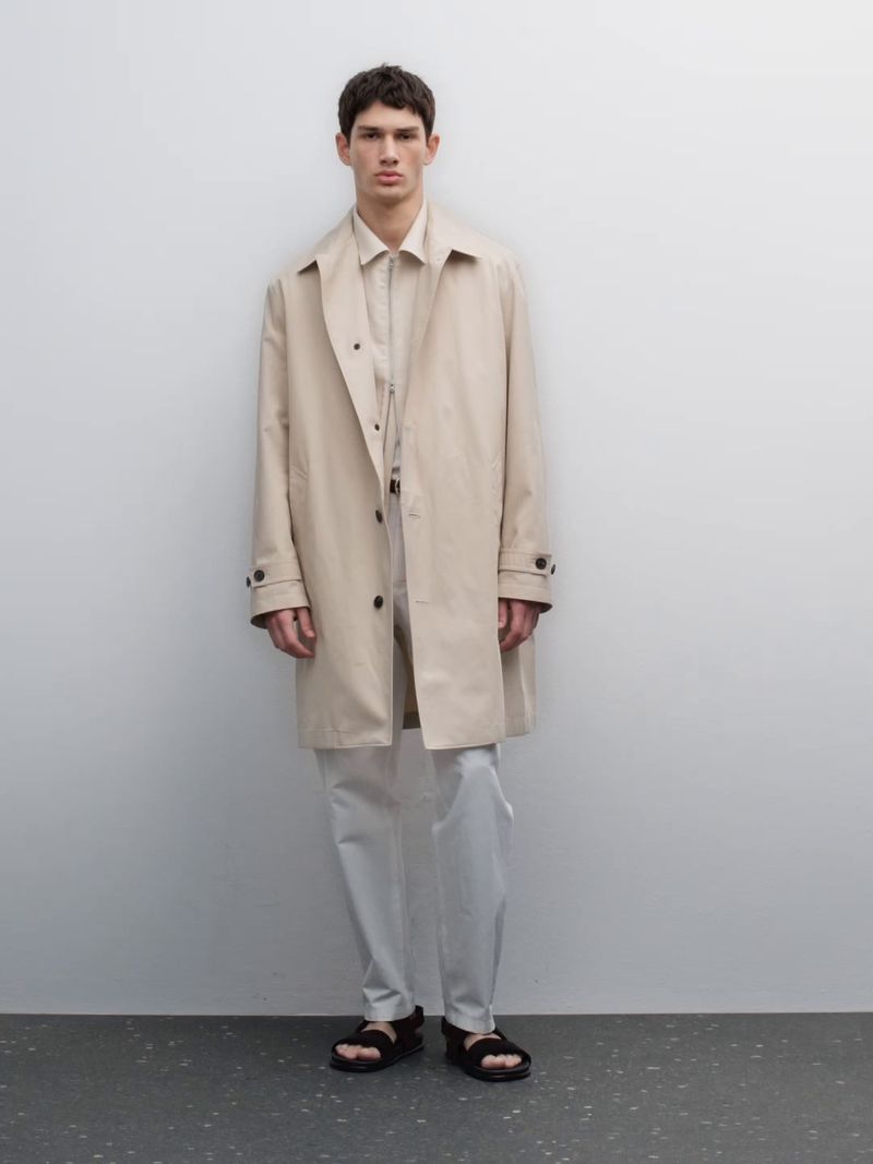 Full-length portrait of a male model wearing a light beige coat and trousers against a neutral grey studio wall.