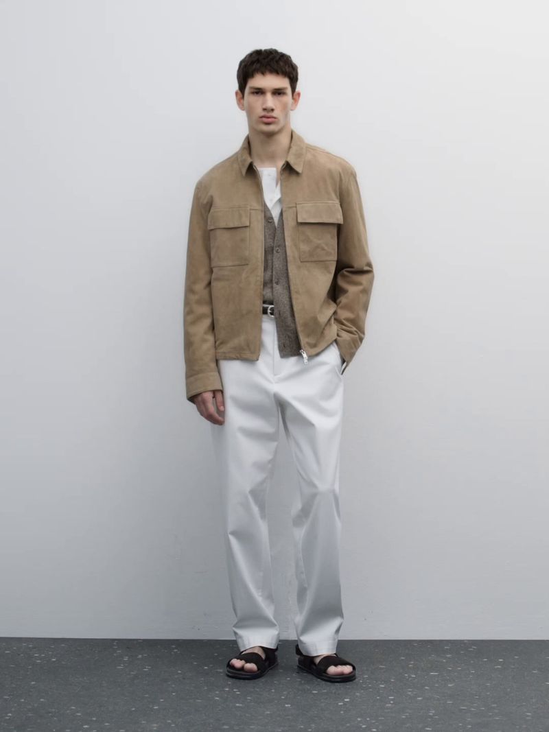 Full-length shot of an adult male model in a beige jacket, vest, white pants, and black sandals, posing against a neutral wall indoors.