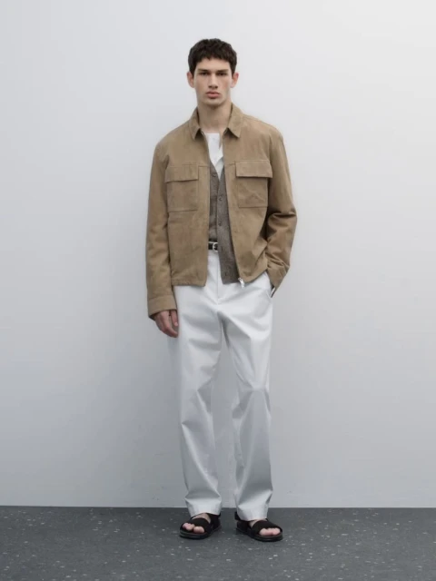 Full-length shot of an adult male model in a beige jacket, vest, white pants, and black sandals, posing against a neutral wall indoors.