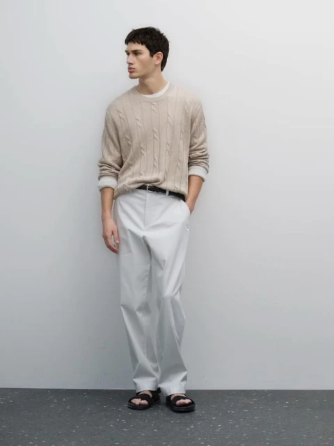 A male fashion model poses in a beige cable knit sweater and white pants against a light background, showcasing a relaxed, neutral aesthetic.
