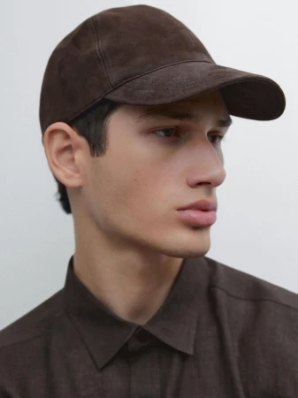Portrait of a young man wearing a brown suede cap and dark shirt against a seamless white background.