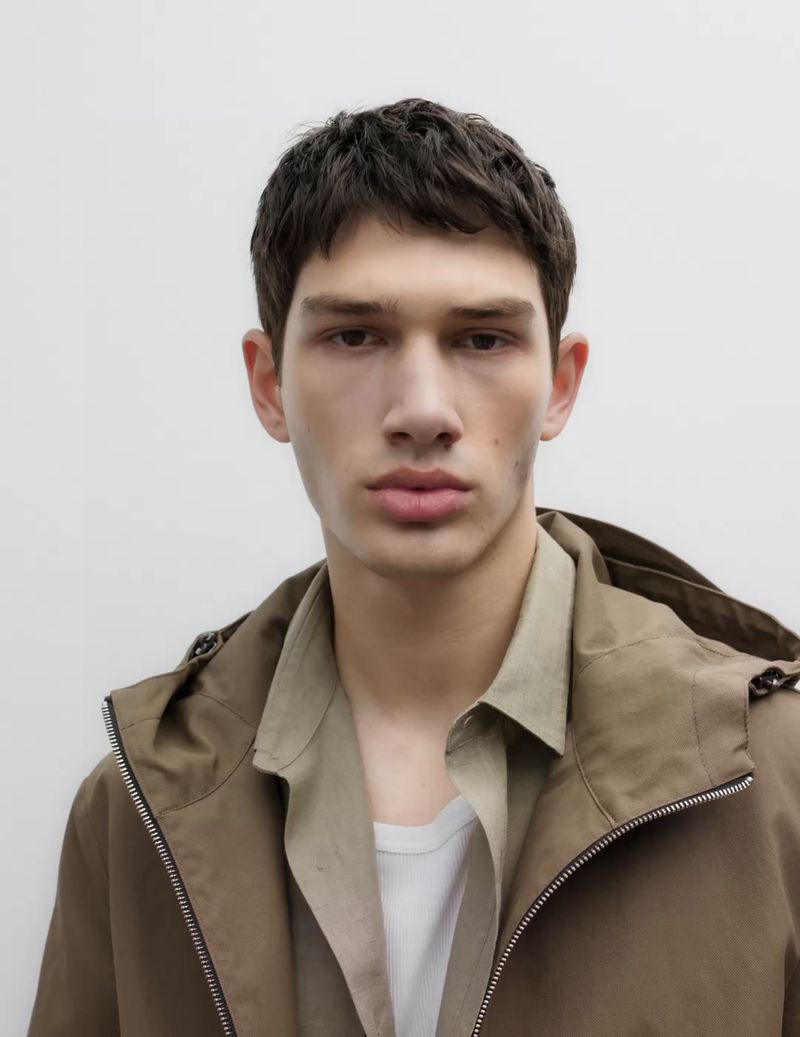 Close-up portrait of a young adult male with short brown hair wearing a brown hooded jacket and a tan shirt against a white background.